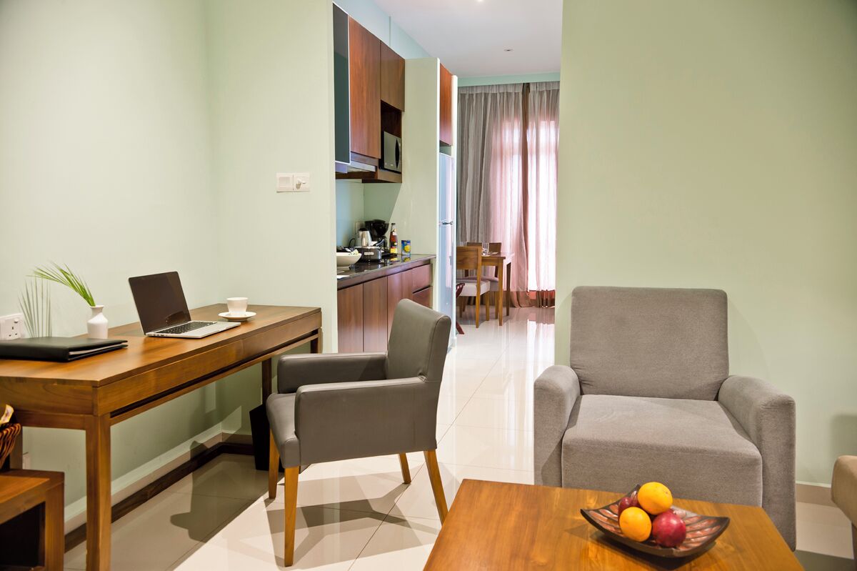 A modern living room with a wooden desk, a laptop, a grey armchair, and a dining area with a table and chairs.