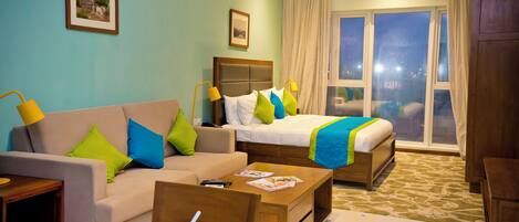 A hotel room with a sofa, a bed with a blue and green bedspread, a wooden coffee table with a remote control and magazines, and a window with curtains.
