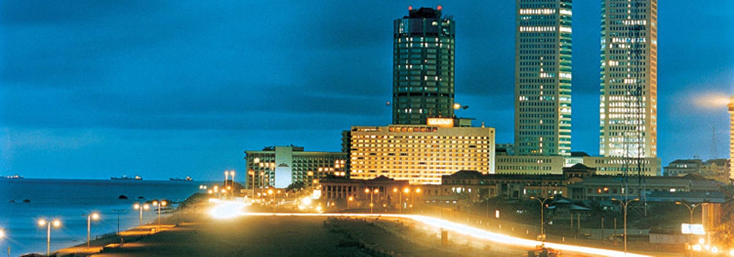 A coastal cityscape at night with high-rise buildings, a beach, and a road with vehicle headlights.