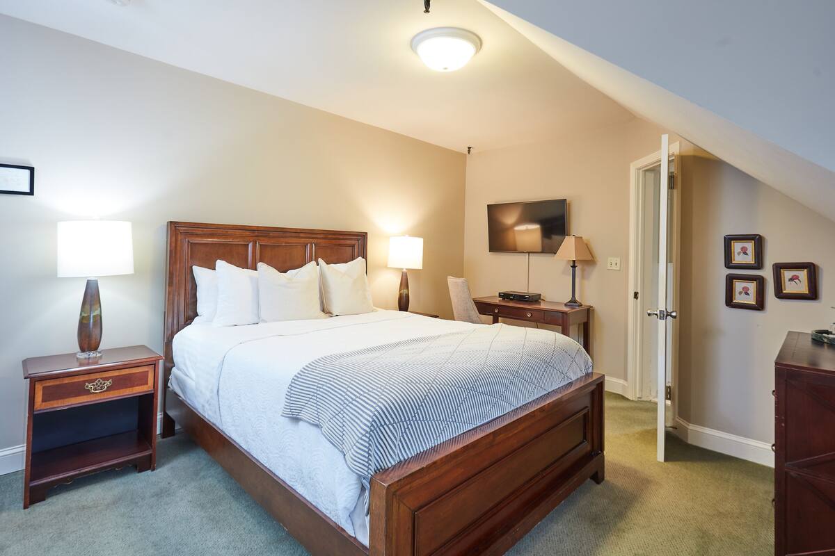 A bedroom with a wooden bed, bedside tables with lamps, a flat-screen TV on the wall, and a door leading to another room.