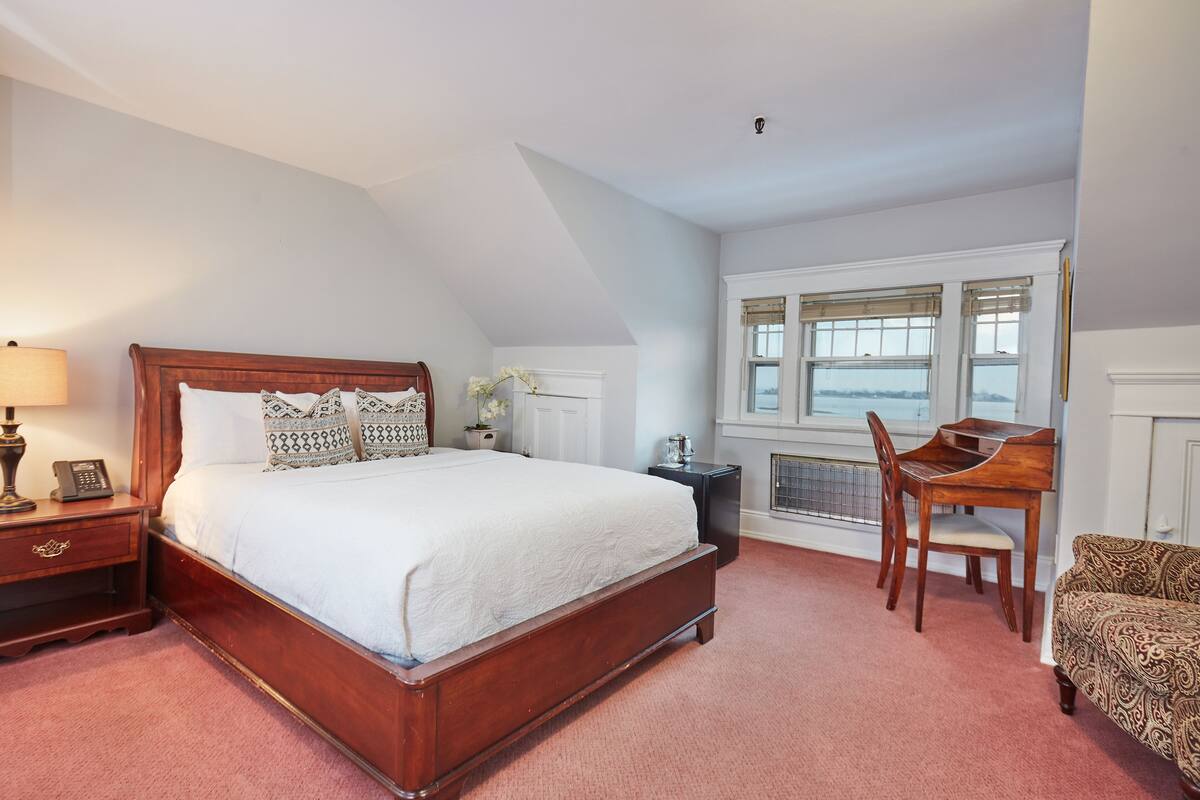 A bedroom with a wooden bed, bedside table with a lamp, a chair, and a window with blinds.