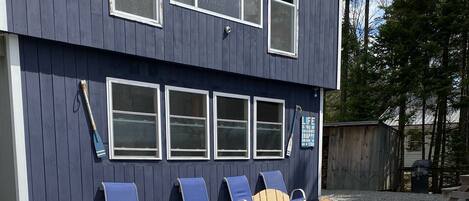 A building with a dark exterior, multiple windows, and a gravel patio with Adirondack chairs and a fire pit.