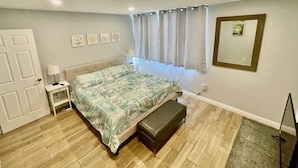 Een slaapkamer met een bed, een bankje, een nachtkastje, een lamp, een raam met gordijnen en een lijst met een afbeelding aan de muur.