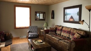 Ein gemütliches Wohnzimmer mit Holzboden, einem großen Fenster, einem Sofa, einem Sessel und einem Couchtisch.