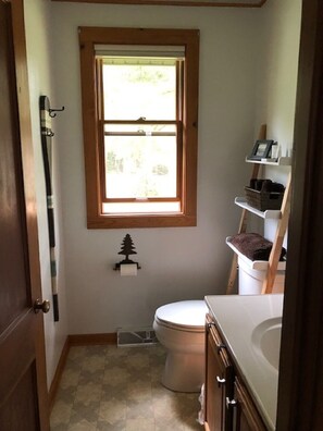 A small bathroom with a toilet, a window, a towel rack, and a shelf with toiletries.