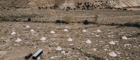 An aerial view of a desert landscape with multiple tents arranged in a field, a vehicle parked nearby, and a large hill in the background.