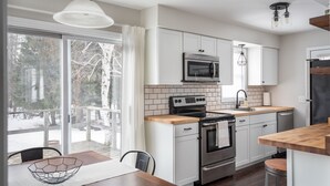 A modern kitchen with white cabinets, stainless steel appliances, and a wooden dining table. - The Elm Cottage - 1mi to Frankfort Beach (Frankfort)