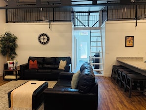 A modern living room with a black leather sofa, a coffee table, and a large potted plant. There is a staircase leading to an upper level.