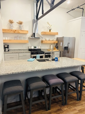 A modern kitchen with a central island, bar stools, stainless steel appliances, and wooden shelves.