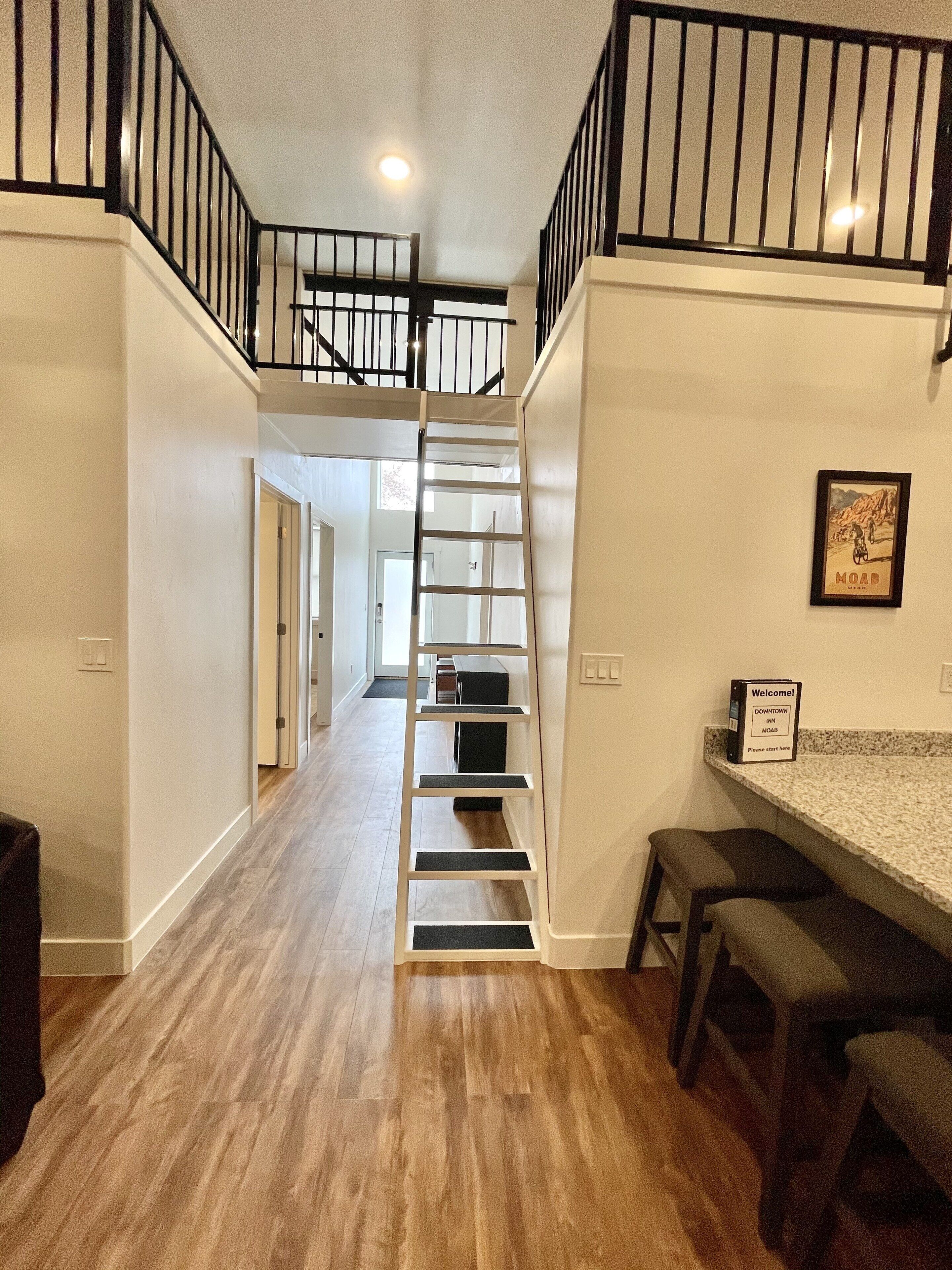 Eine Treppe führt zu einer zweiten Etage mit Metallgeländer, einem bordgestellten Willkommensschild und einer Bar mit Hockern.