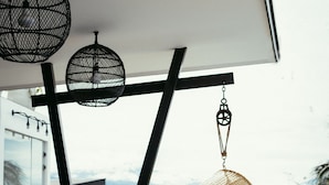 A hanging chair with a woven seat suspended from a ceiling, a table with a round shadow on the floor, and a view of a mountainous landscape through a window.