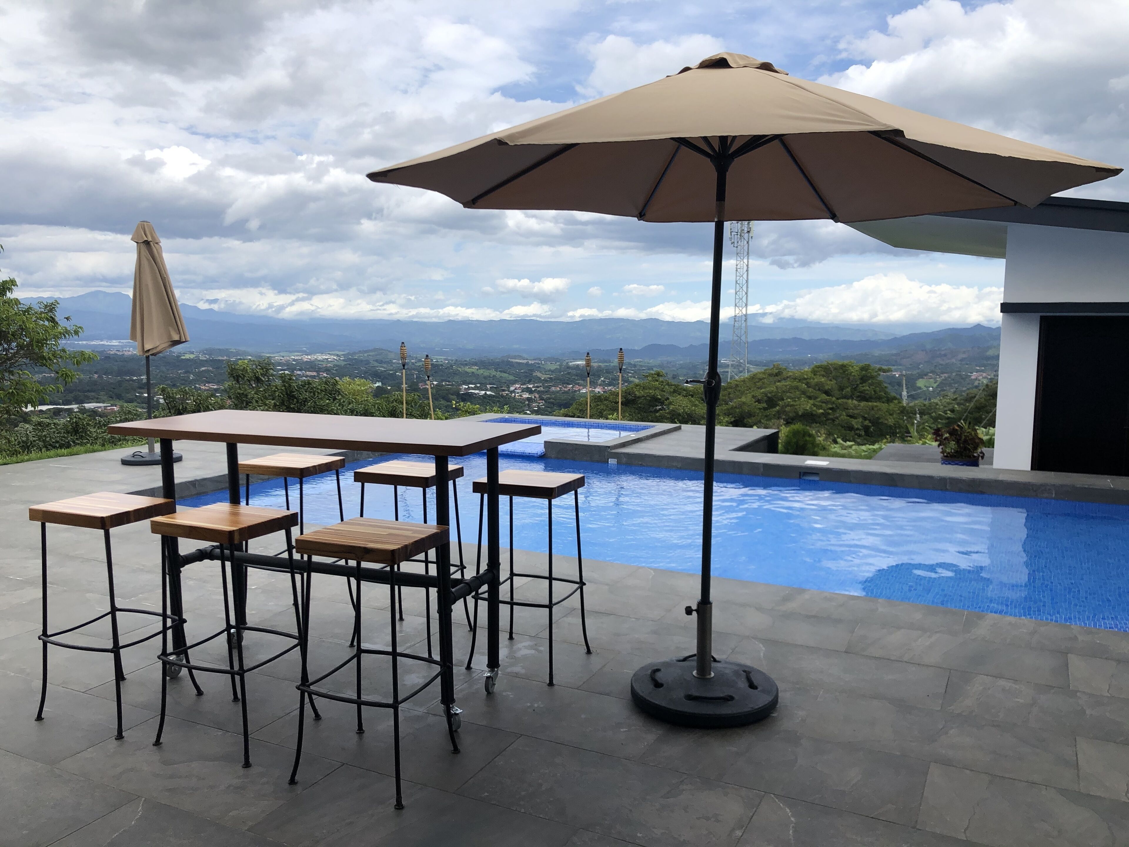An outdoor pool area with a table and chairs, a large umbrella, and a view of mountains in the background.