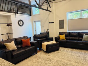 A spacious living area with two leather sofas, a coffee table, and a patterned rug. A clock and a coat rack are visible on the walls.