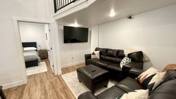 A modern living room with a black leather sofa, a flat-screen TV mounted on the wall, and a wooden floor.