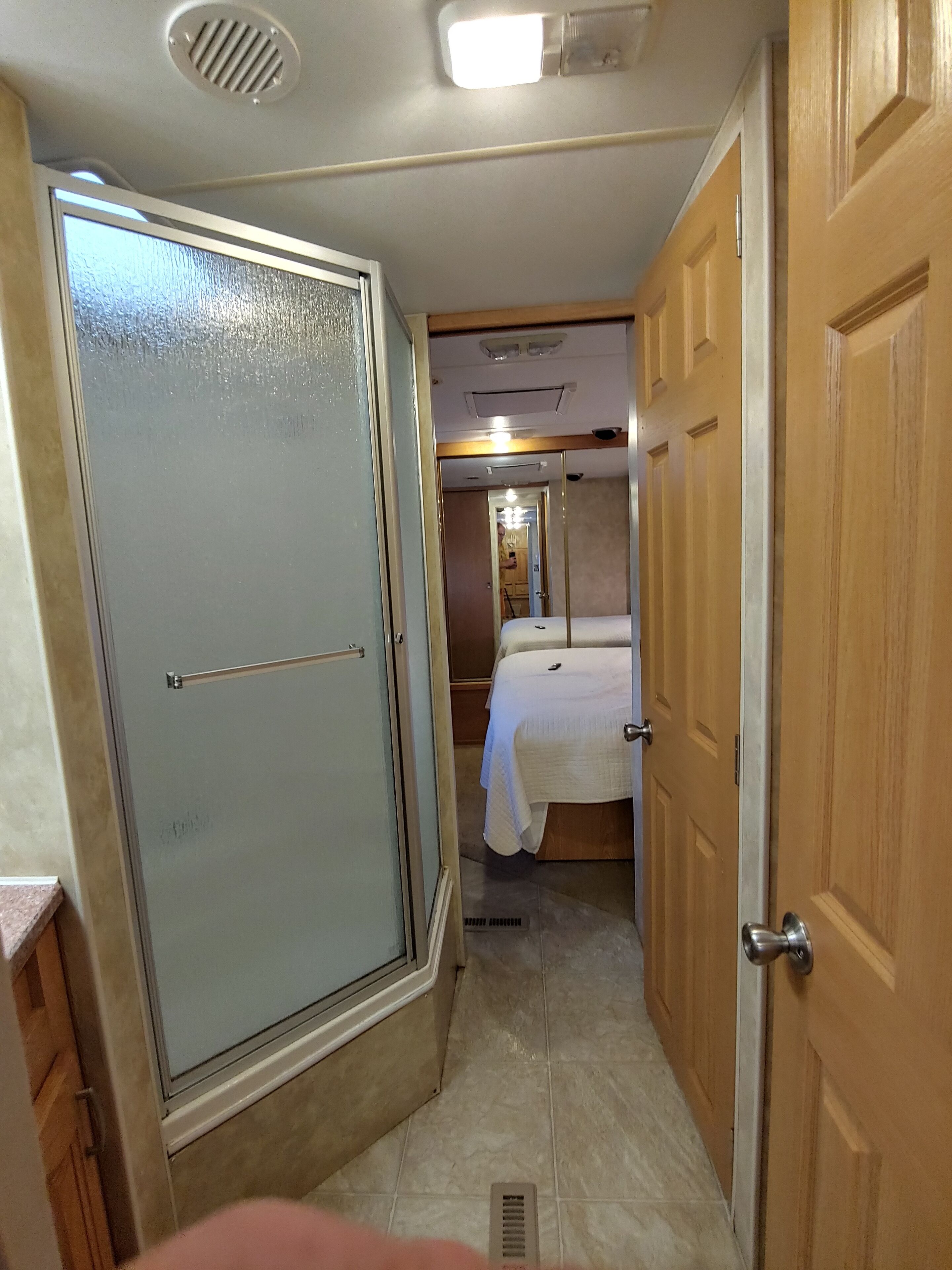 Un estrecho pasillo con una puerta corredera a la izquierda, una cama con colcha blanca en el centro y puertas de madera a la derecha.