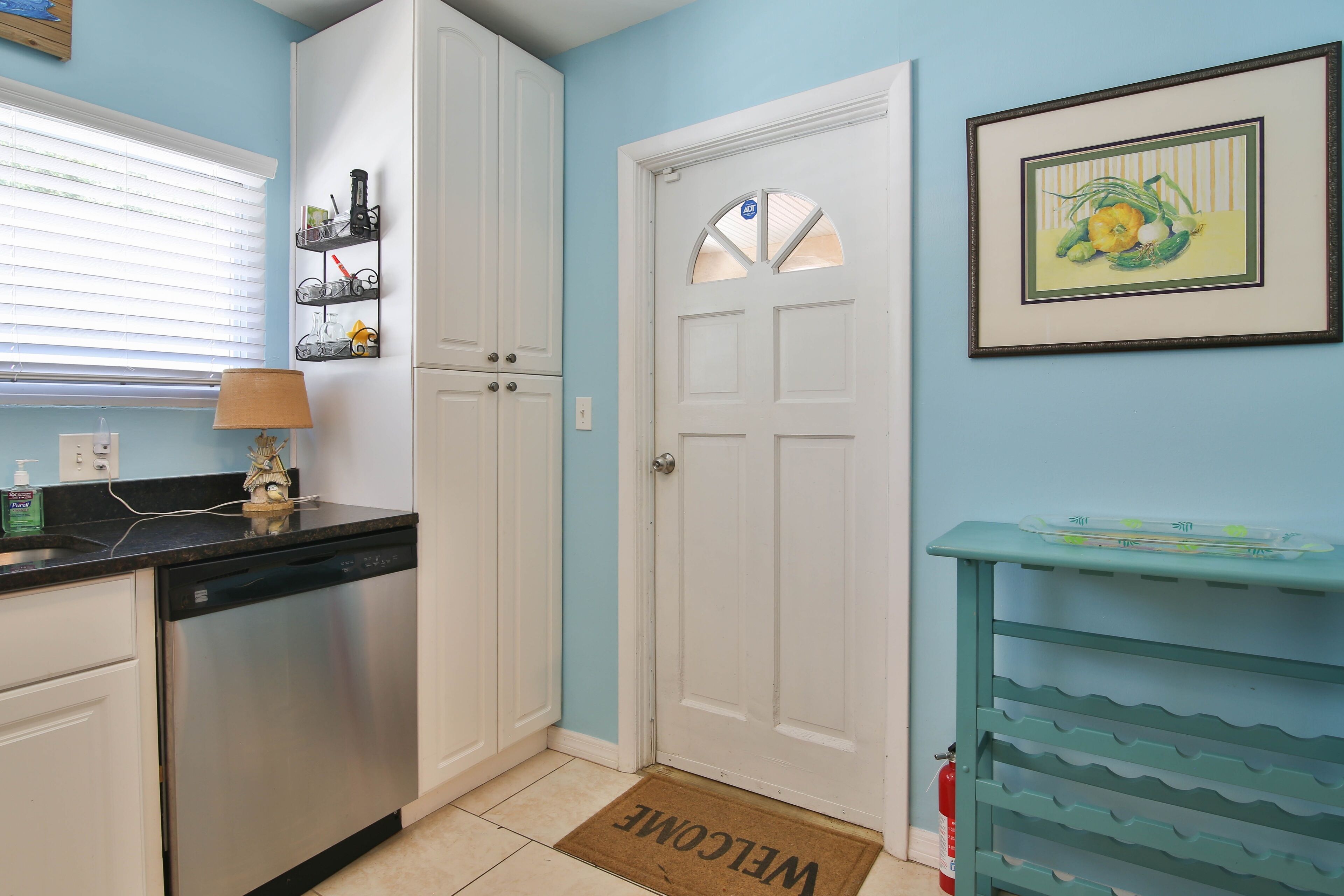 A kitchen with white cabinets, a dishwasher, a welcome mat, a framed picture, and a teal shelf unit.
