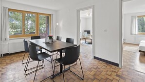 Un comedor moderno con una mesa rectangular, seis sillas negras y un jarrón con flores. Se observan amplias ventanas con cortinas, un radiador y una lámpara en el techo.