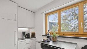 Una cocina moderna con armarios blancos, lavavajillas integrado, cafetera y ventana con vistas a árboles y montañas.
