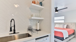 A modern kitchen with a stainless steel sink, white tiled backsplash, and a dishwasher. There are white shelves above the sink with various items.