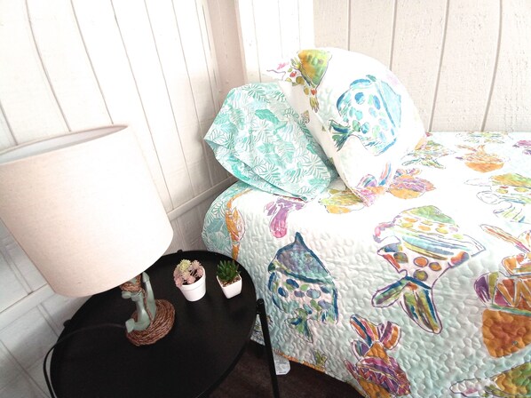 A bedroom with a bed covered in a colorful quilt, a bedside table with a lamp and small potted plants, and a white wooden wall.