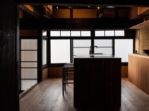 A modern kitchen with a central island, a bar stool, and a large window with blinds. - Maana Kamo (Kyoto)