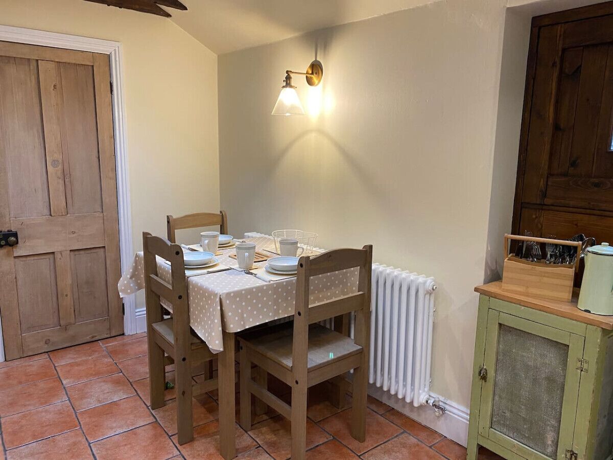 Comedor con una mesa dispuesta para cuatro, sillas de madera, una lámpara fijada a la pared, un radiador y un armario de madera con encimera clara.