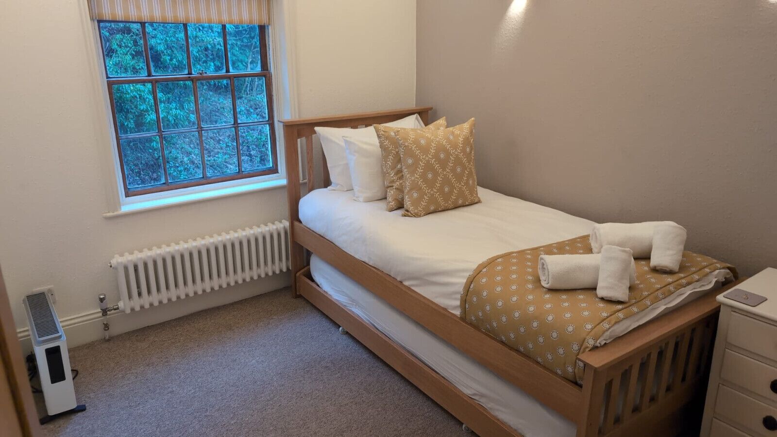 A neatly made bed with a white and patterned bedspread, a radiator below, and a window with a view of greenery.