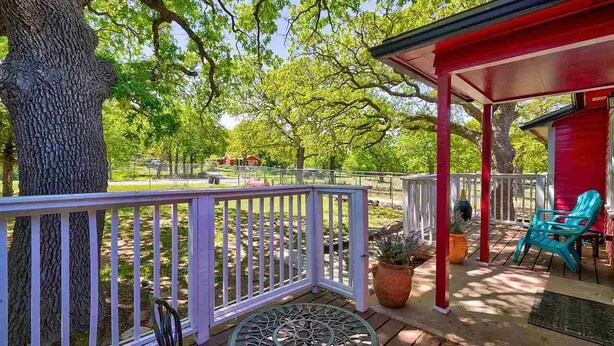 Een overdekt terras met een tafel en stoelen, omgeven door bomen en een rood gebouw.