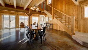 A spacious room with a high ceiling, wooden beams, a large dining table with chairs, a staircase, and large windows offering a view of the mountains. - Sunset Grove: A Yellowstone Retreat (Livingston)