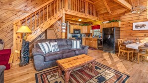 Interior acogedor de una cabaña con sala de estar, cocina y comedor, que presenta pisos de madera, una escalera y un ventanal grande.