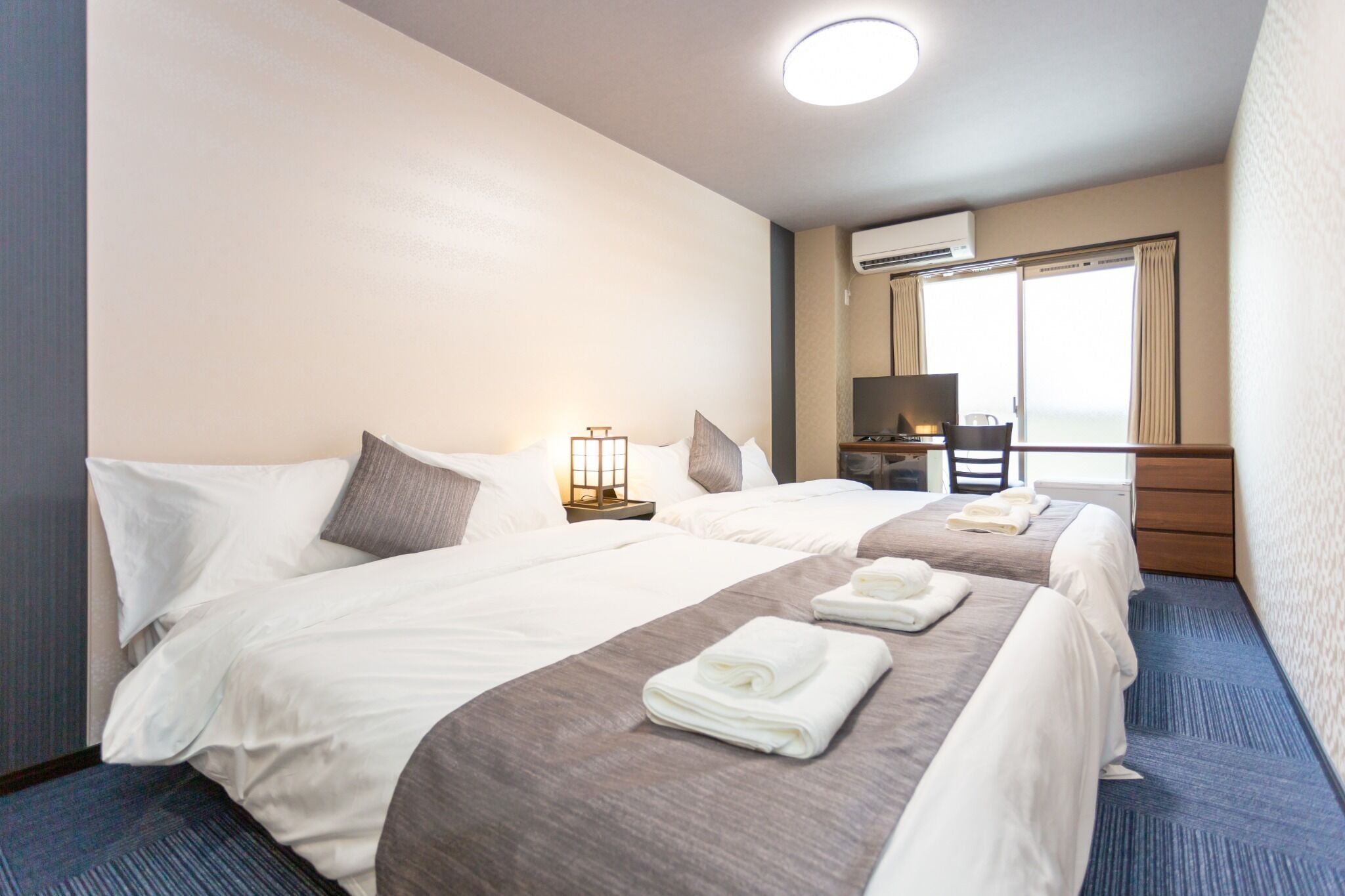 A hotel room with two beds, a desk, an air conditioning unit, and a large window with curtains.
