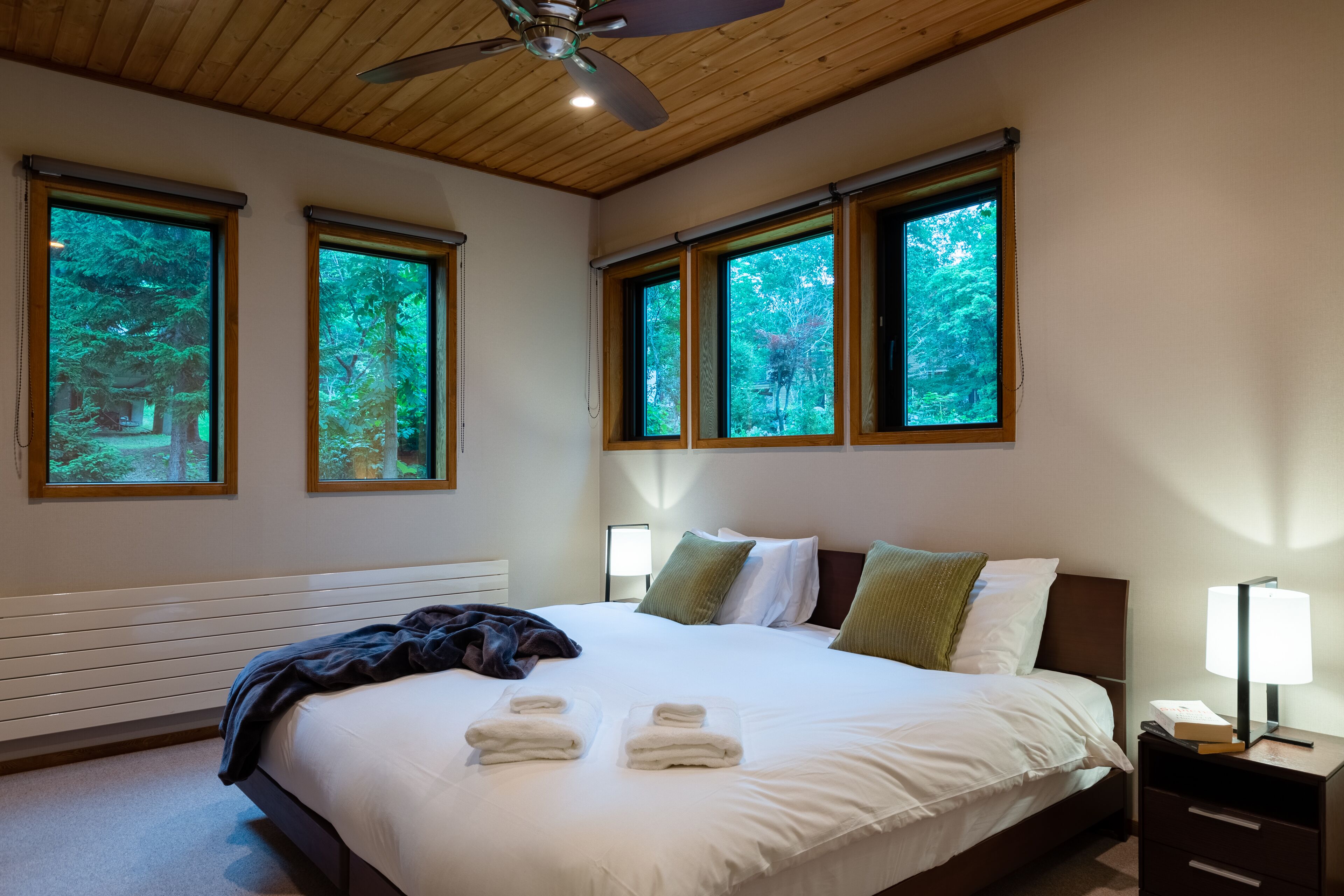 A bedroom with a large bed, two windows with a forest view, a ceiling fan, and a bedside table with a lamp.