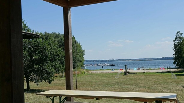 Un pabellón exterior con mesas de picnic con vistas a un lago con barcos y una playa.