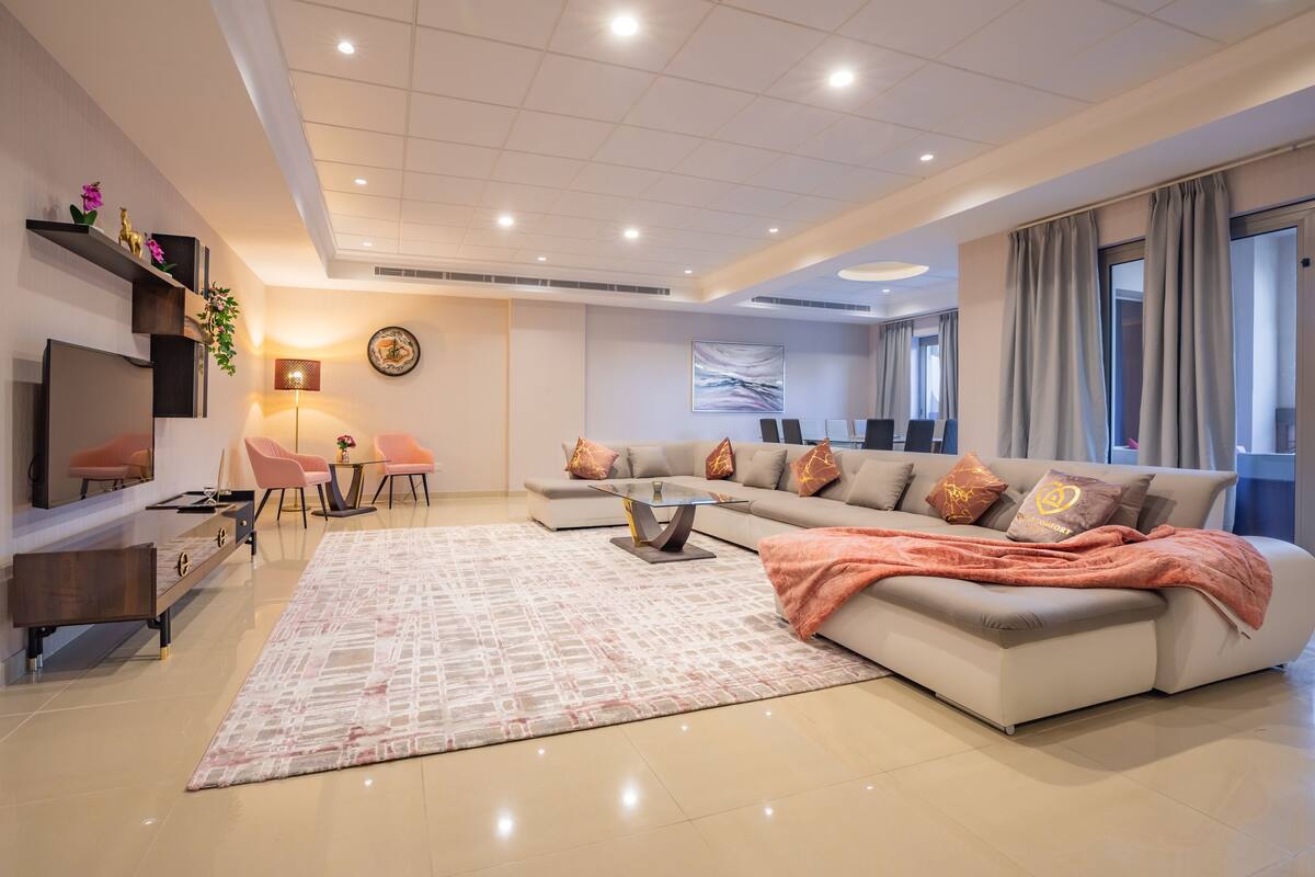 A modern living room with a sofa, coffee table, and TV. There is a dining area with chairs and a table. The room has a large window and a ceiling with recessed lighting.
