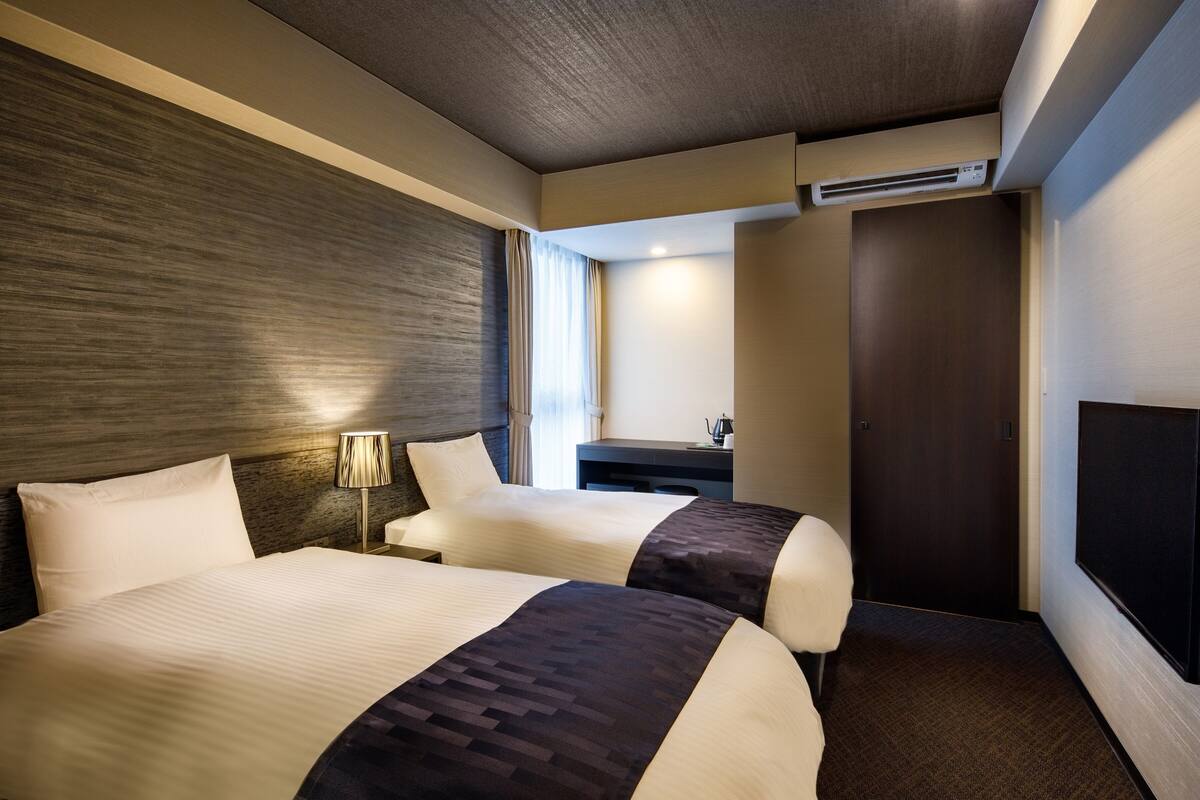 A hotel room with two beds, a desk, a chair, a lamp, and a TV.