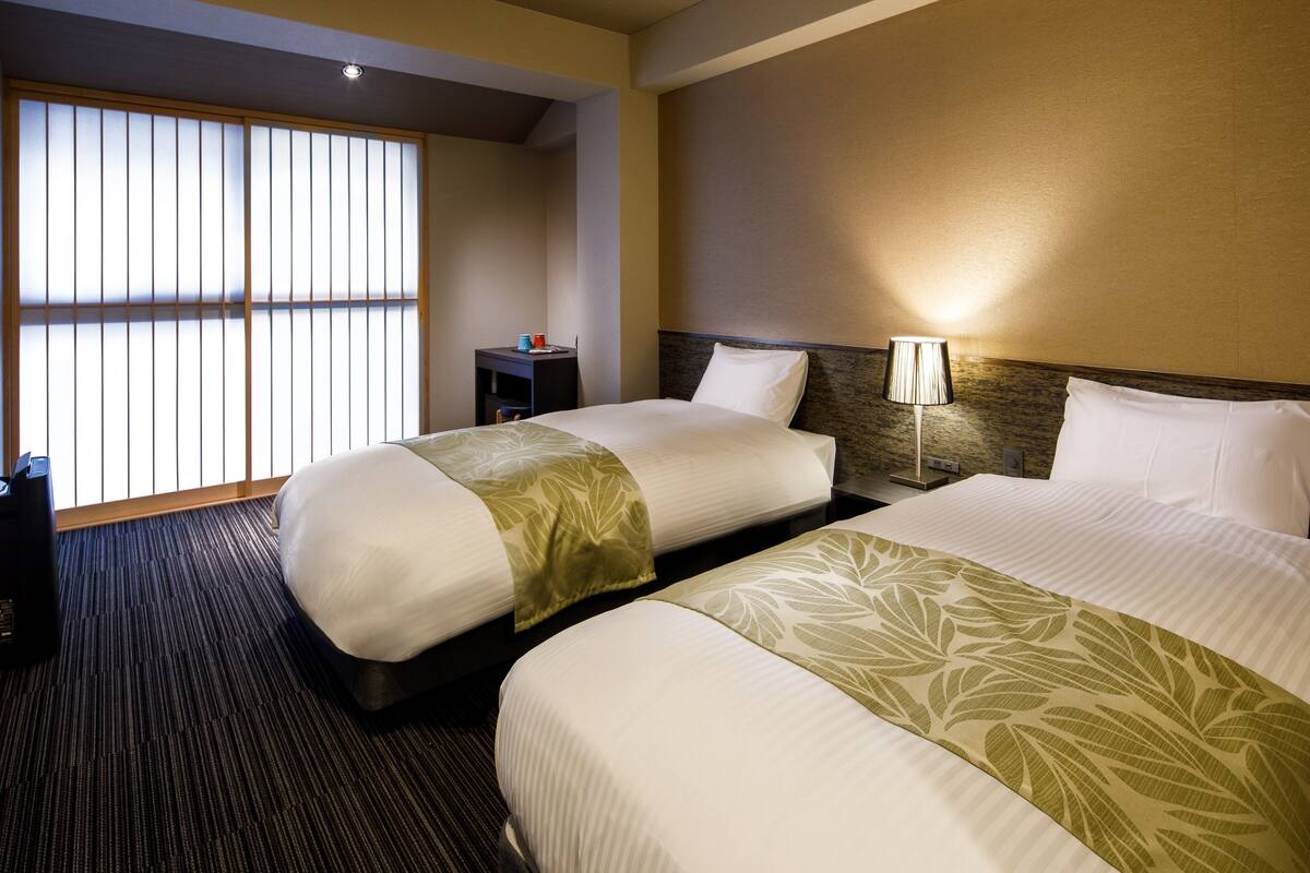 A hotel room with two beds, a nightstand, a lamp, and a window with blinds.