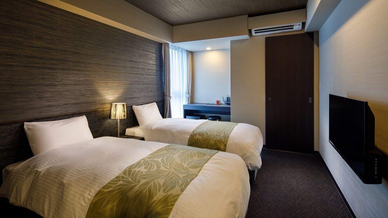 A hotel room with two beds, a desk, a TV, and an air conditioning unit.
