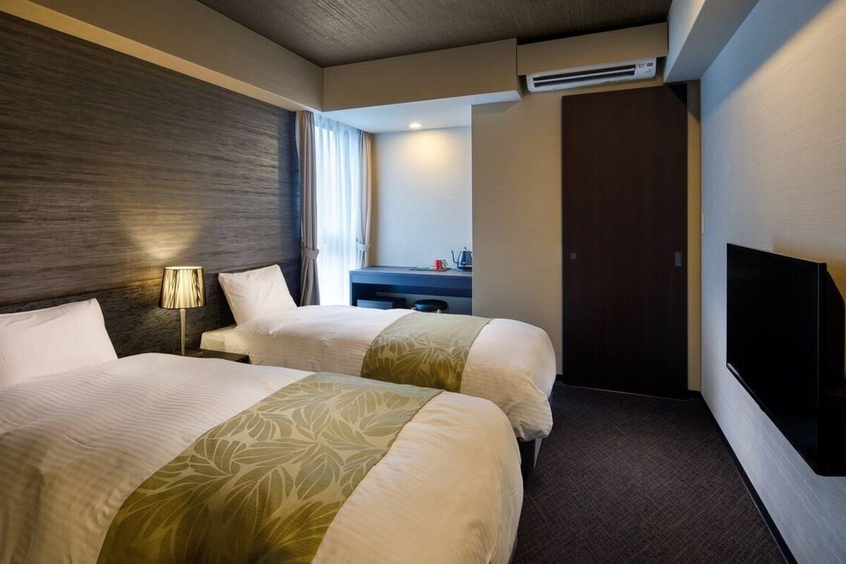 A hotel room with two beds, a desk, a TV, and an air conditioning unit.