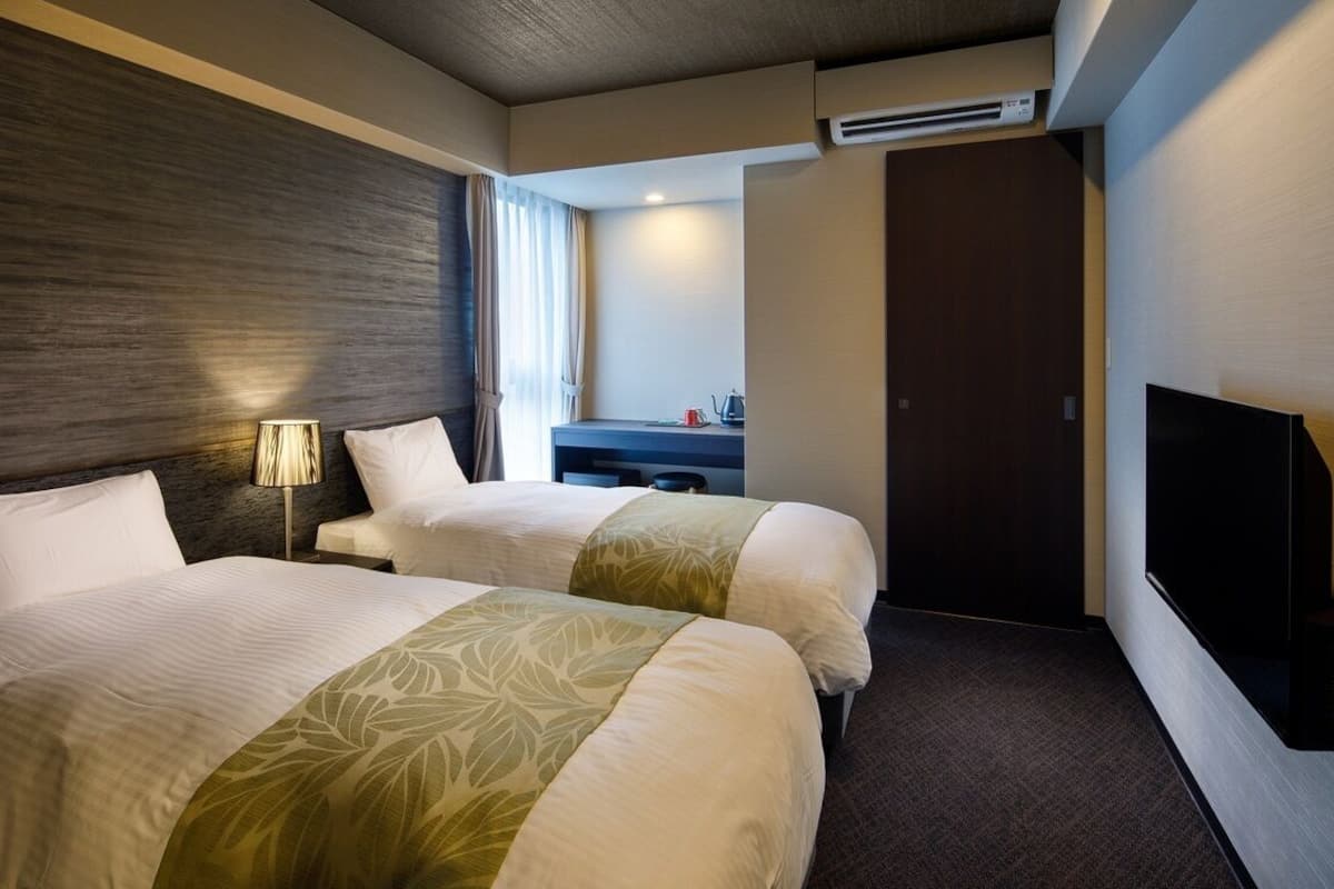 Ein Hotelzimmer mit zwei Betten, einem Schreibtisch, einem Fernseher und einer Klimaanlage.