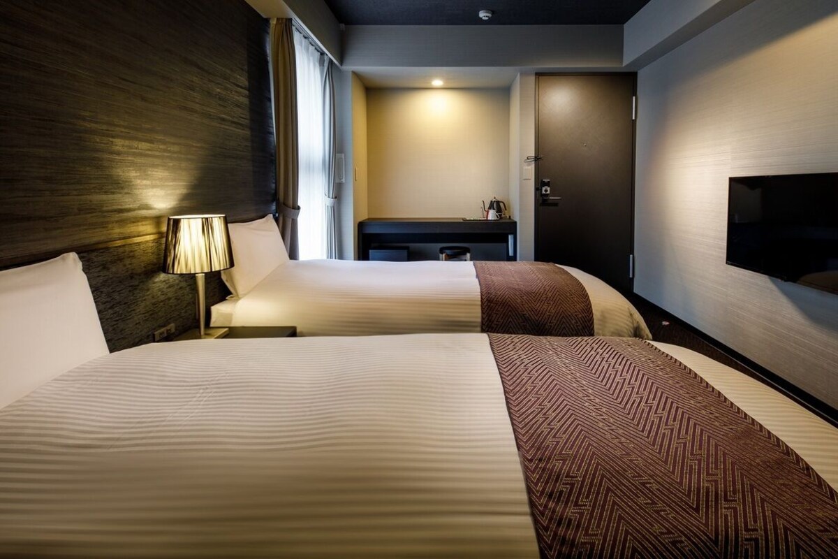 A hotel room with two beds, a desk, a TV, and a door.