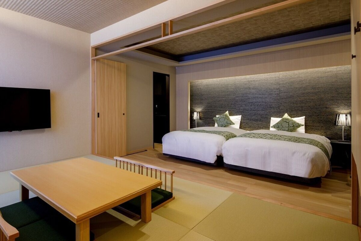 A modern hotel room with a flat-screen TV, a wooden table, two beds with green bedding, and a built-in headboard.