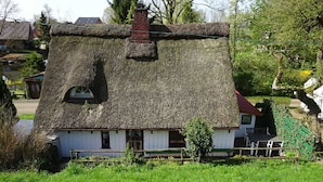 Une maison traditionnelle à toit de chaume, dotée d’une petite fenêtre et d’une cheminée, entourée d’un champ verdoyant et luxuriant ainsi que d’arbres.