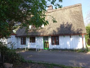 Una casa tradicional con techo de paja, paredes blancas y detalles verdes, con un banco en el exterior y una puerta con marco verde.