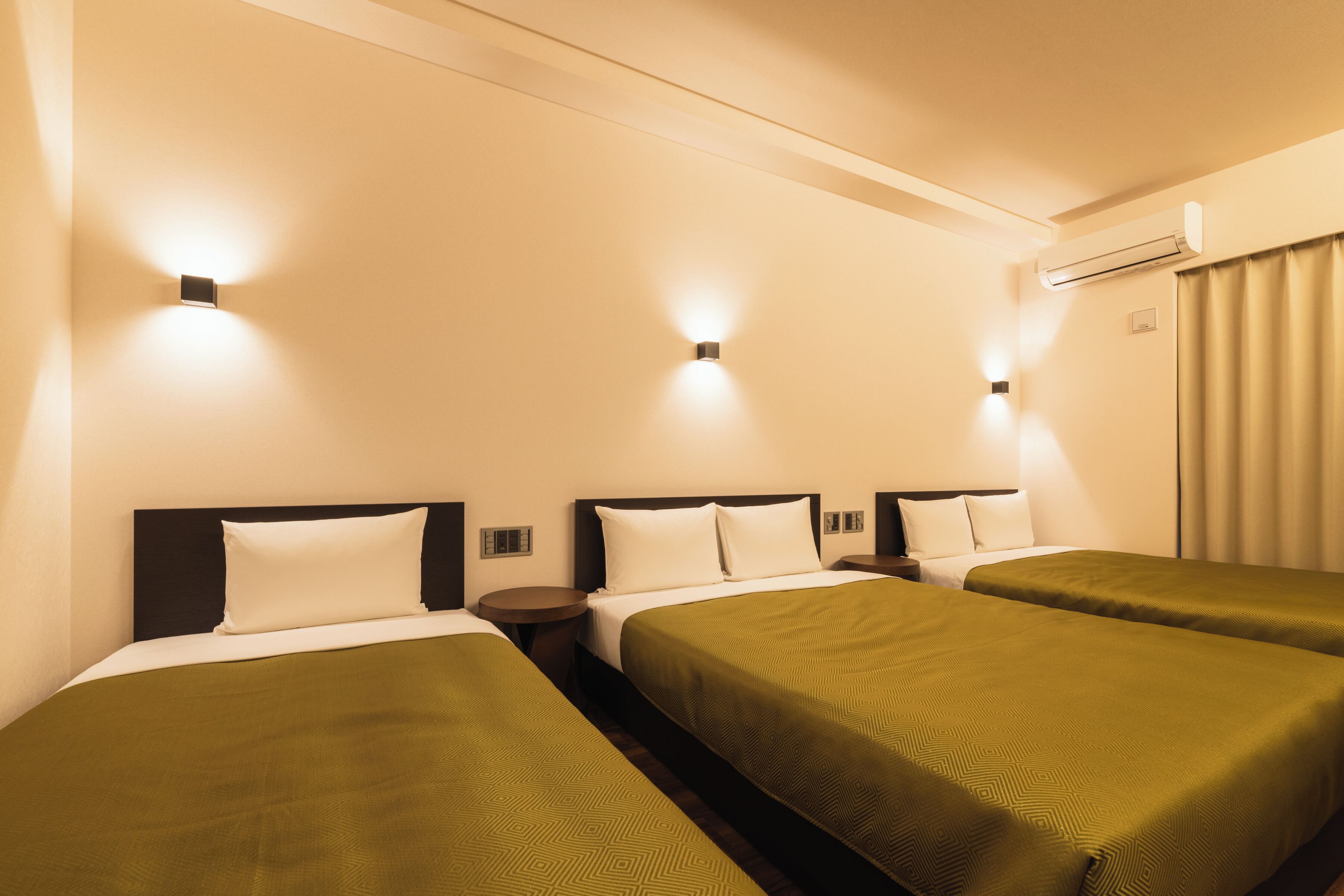 A hotel room with two single beds, a nightstand, a wall-mounted air conditioner, and a window with curtains.