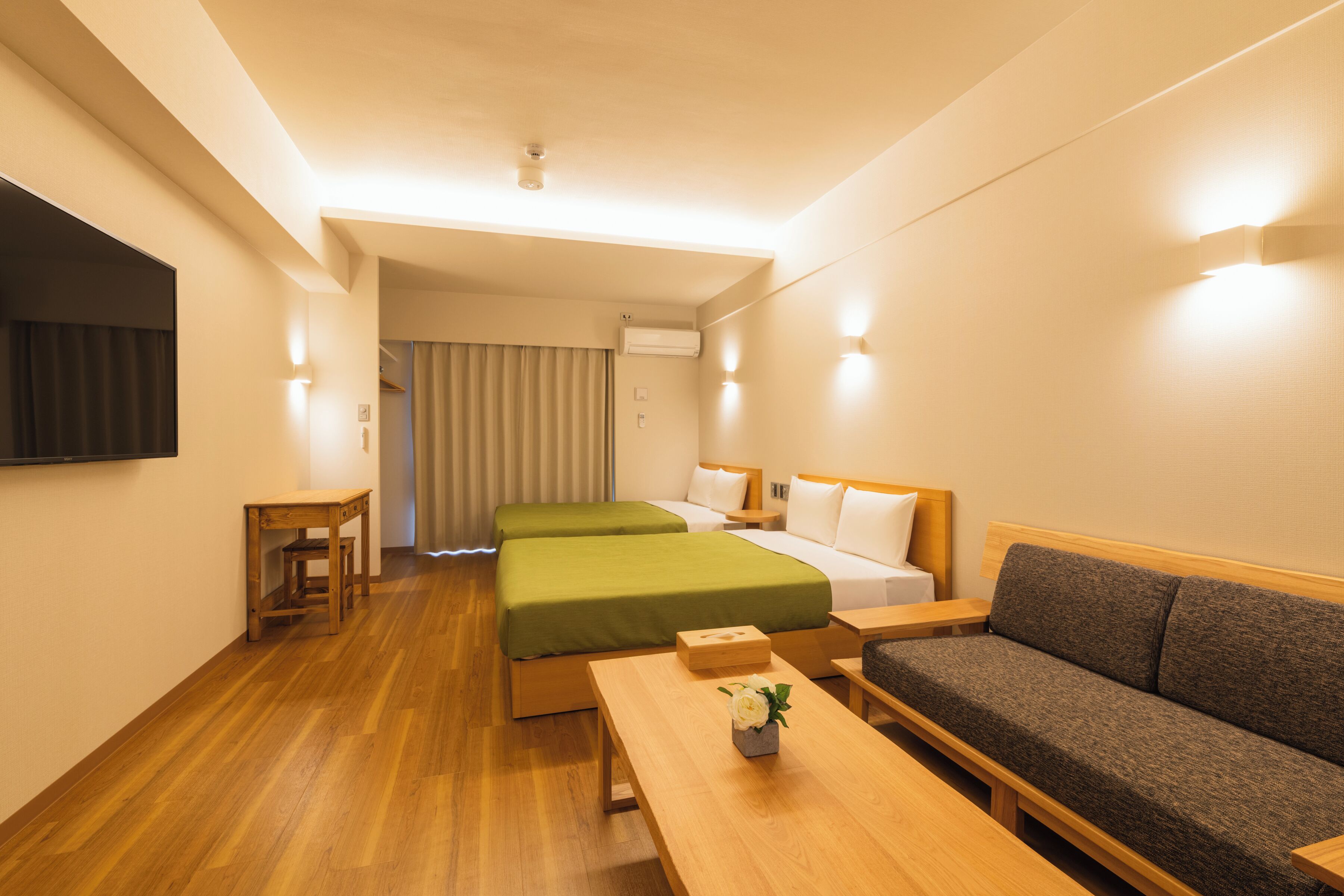 A hotel room with two beds, a sofa, a wooden table, and a TV mounted on the wall.