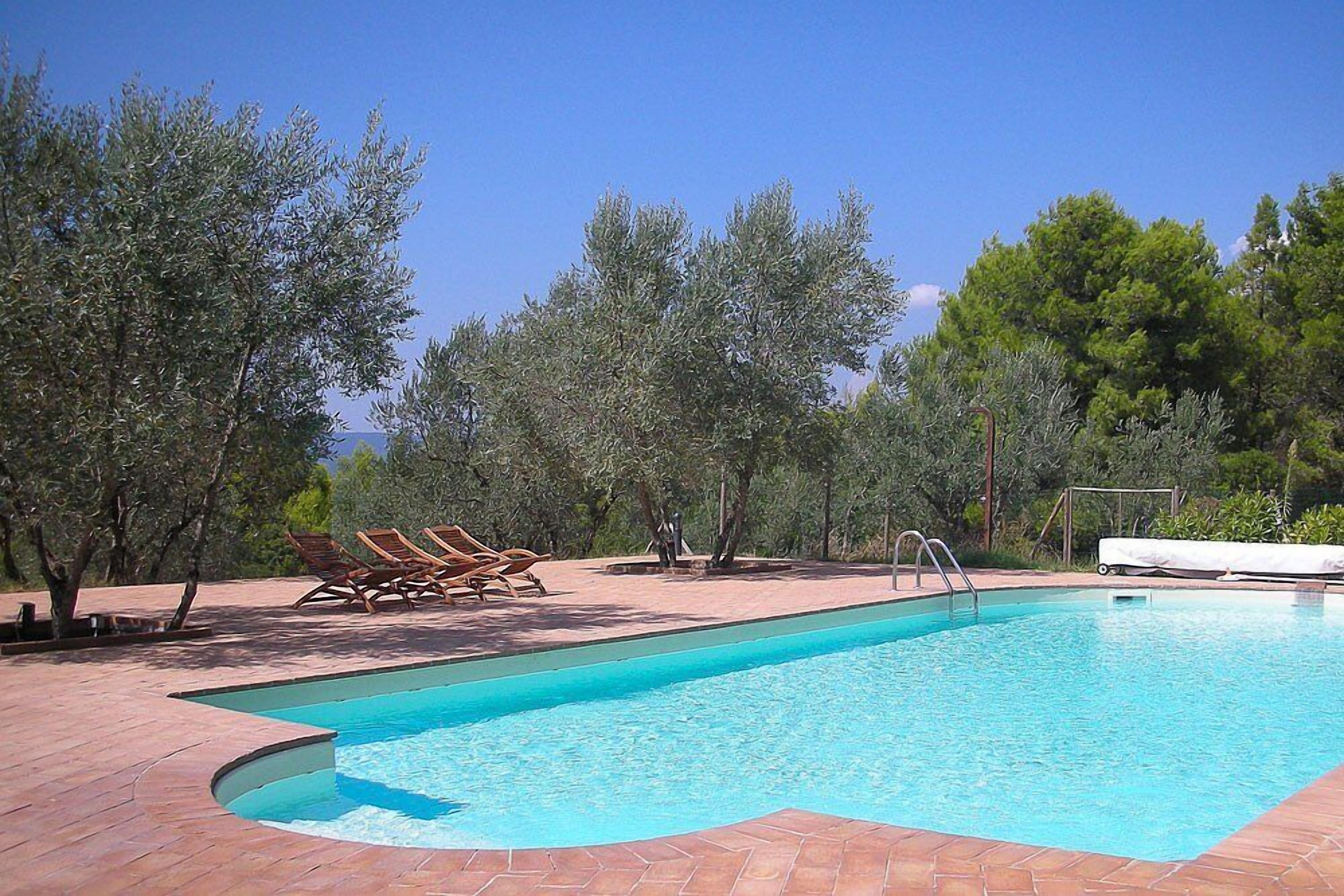 A rectangular swimming pool with a tiled edge, surrounded by a brick patio. There are lounge chairs and a small white table near the pool.