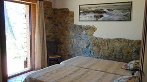 Een slaapkamer met een stenen muur, een bed met bloemenkussens, een stoel en een schilderij van een zeegezicht.
