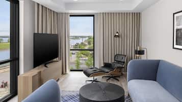A modern hotel room with a large window offering a view of a bridge and greenery, a flat-screen TV mounted on a wooden console, a blue sofa, a black leather chair, and a small round table.