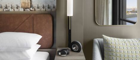A hotel room with a bed, bedside table with headphones, a lamp, a mirror, and a view of a bridge.
