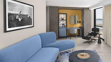 A modern hotel room with a blue sofa, a small round table, a desk with a chair, and a large window offering a view of a bridge.